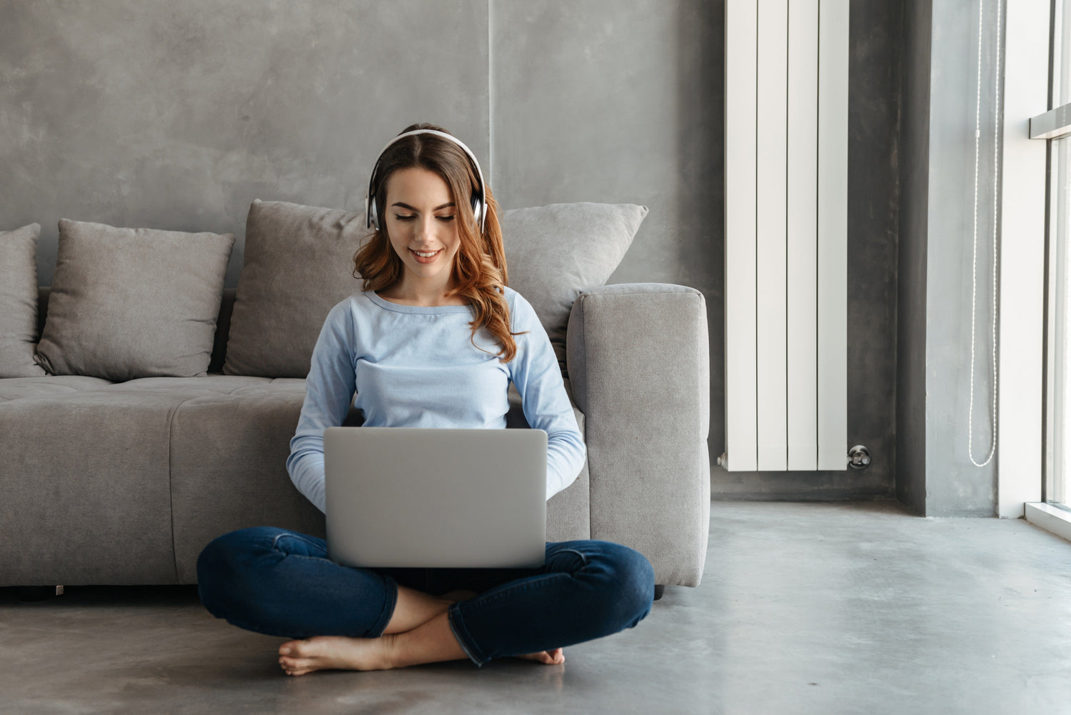 Junge Dame im Schneidersitz auf dem Boden eines modernen Wohnzimmers, trägt Kopfhörer und nutzt einen Laptop, der auf ihrem Schoß liegt